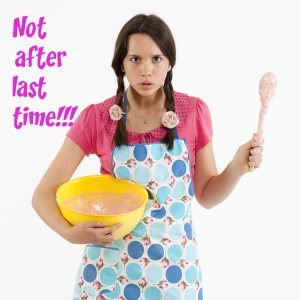 An angry teenage girl dressed for baking waves her wooden spoon like a weapon.