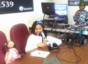 A little girl DJ sits next to her Zolie Zoi doll while preparing to read a story on air.